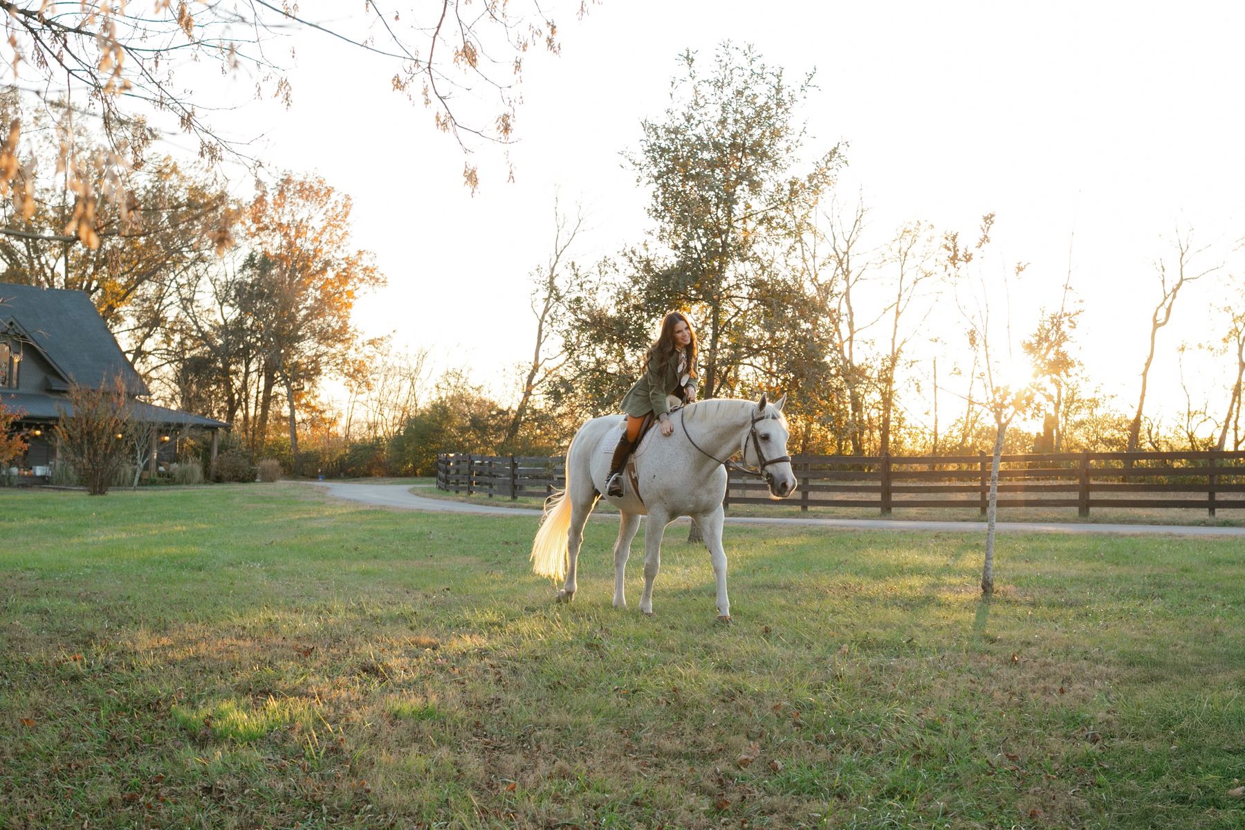 Tiffany riding a white horse across a pasture at sunset, with a farmhouse and black board fence behind her.
