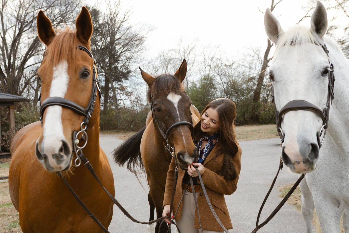 Tiffany smiling at a bay horse between a chestnut and a gray horse on the farm lane.